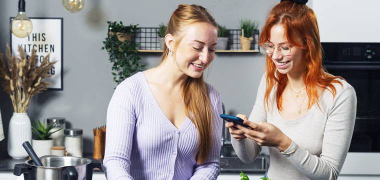 Isy und Tessa stehen hinter einer Arbeitsfläche und schauen auf ein Handy.